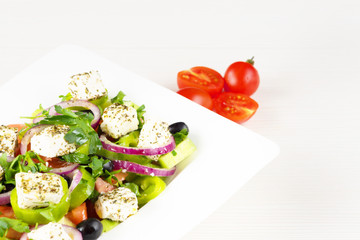 Greek salad with fresh tomato, cucumber, red onion, basil, feta cheese, black olives, Italian herbs and olive oil in white dish on white wooden table.