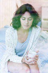 Young beautiful brunette woman sitting down home on the bed by the window with cup of coffee