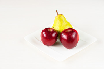 Red apples, green pear in white plate on white wooden background.