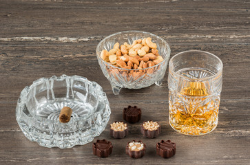 Glass of whiskey, a cuban cigar, nuts and chocolates on a grey wooden table. Close-up view.