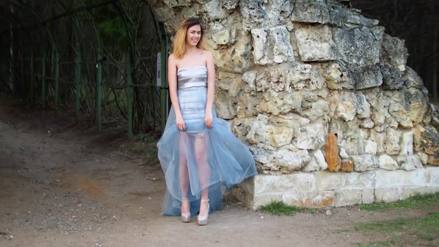 Attractive girl with black brows and curly hair in silver and blue dress approaches camera showing legs in high heeled shoes during photoshoot.