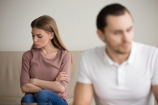 Upset disappointed girlfriend is angry at boyfriend after fight and waiting for apology, young offended woman sitting with arms crossed sulking, unhappy couple after quarrel sitting apart on couch