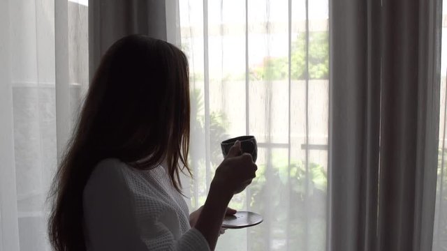 Young woman with long hair in white bathrobe drink coffee standing near the window at home.