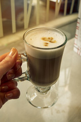 Hand holding Glass of Tasty classic coffee latte macchiato with foam. Caf&eacute; or restaurant. Closeup. Toning.
