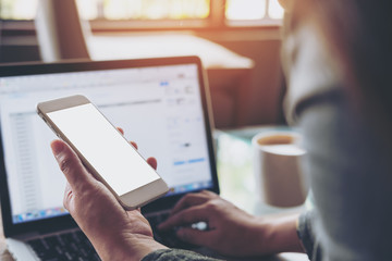 Mockup image of a woman holding mobile phone with blank white screen while using laptop with coffee cup on the table in office