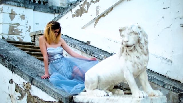 Attractive girl with black brows and curly hair in silver and blue dress sits on stone balustrade near lion statue and poses during photoshoot in antique estate.