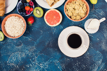 Top view view of healthy breakfast on the blue concrete table, coffee with pancakes with berries, cereals, oats, banana, grapes, croissant, nuts, selective focus. Copy space for text, square