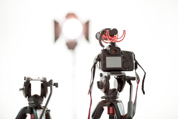 A digital video camera with a microphone on a tripod on a white background, a bright spotlight in the background.