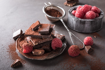 Broken chocolate pieces and raspberries with cocoa powder on metal plate on brown background with copy space. Dark photo.