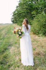 Fototapeta na wymiar bouquet, people, flower holidays , gift and floral arrangement concept - beautiful female in a fashionable white dress posing with her back with bouquet of fresh flowers on background of trees