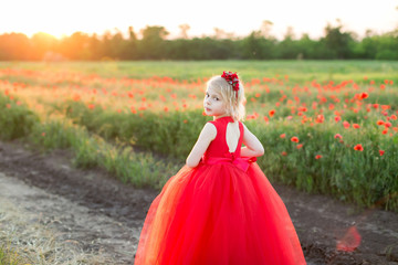Fototapeta premium girl model, childhood, fashion and summer concept - girl blonde in red dress standing half a turn on the road near the field with poppies, on the back of her dress neckline, on head a red headband
