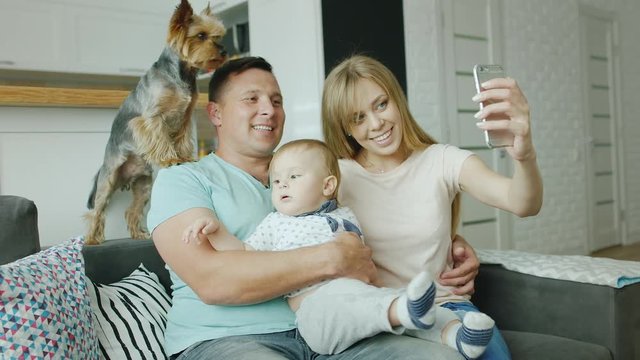 A young family - father, mother, little son and a dog do selfie at home. They sit on the couch, smile at the camera, take pictures with the phone. Happy family
