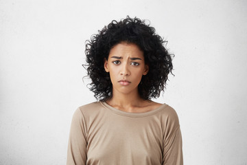 Frustrated young dark-skinned woman with shaggy black hair frowning, looking at camera with sad and...