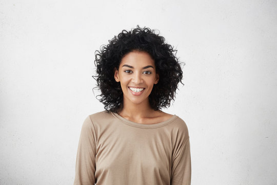 Waist up portrait of casually dressed young mixed race female with curly hair smiling cheerfully during audition for role in TV series, feeling excited and a big nervous, trying to impress director