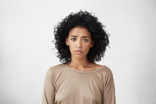 Frustrated young dark-skinned woman with shaggy black hair frowning, looking at camera with sad and unhappy expression while going through hard times in her life. Human emotions and feelings