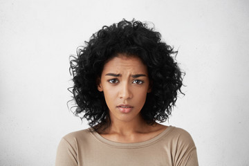Beautiful young dark-skinned woman wearing beige top frowning looking at camera, having clueless...