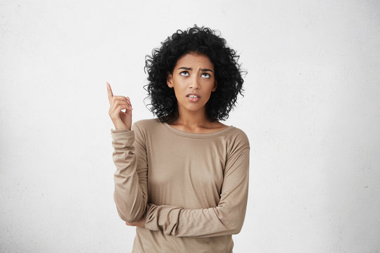 Studio picture of angry and indignant young mixed race female with Afro hairstyle looking up and pointing index finger upwards, feeling mad with noise coming from neighbors above. Body language