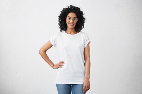 Stylish young dark-skinned female with curly hair looking at camera and smiling cutely, posing with hand on her waist, isolated on white background with copy space for your promotional content