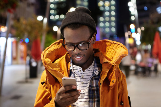 Headshot of handsome young Afro American student having walk around night city, holding mobile phone, using urban wifi, browsing pictures on social media. Modern technology and communication