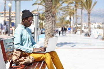 Outdoor summer shot of concentrated young black European traveler with laptop pc sitting on bench on promenade and using free city wi-fi for buying airplane tickets while traveling by himself