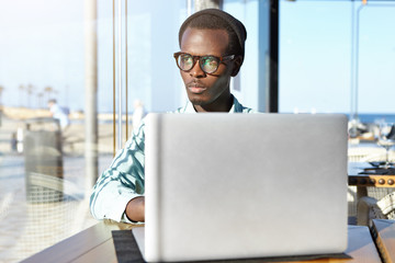 Serious young African Americans programmer working on notebook pc at coworking space, sitting in front of open laptop and looking through window outside, having thoughtful pensive expression