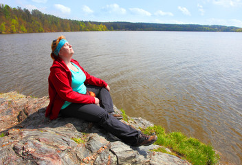 Overweight hiker woman relaxing and meditating on a cliff over lake. Active lifestyle and mental health theme.