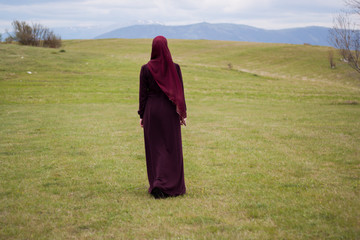 Young and beautiful girl covered, Muslim, in burgundy dress, walk through the meadows surrounding hills, enjoying the natural resources, relaxation