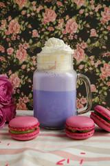 Purple coffee in stylized mason jar cup with macarons and roses and colorful decoration on background of floral pattern. Blueberry milk shake. Unicorn coffee. Unicorn food