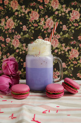 Purple coffee in stylized mason jar cup with macarons and roses and colorful decoration on background of floral pattern. Blueberry milk shake. Unicorn coffee. Unicorn food