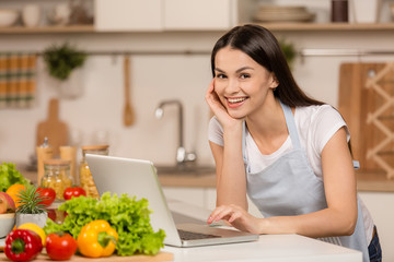 Obraz na płótnie Canvas Young Woman in kitchen with laptop computer looking recipes, smiling. Food blogger concept