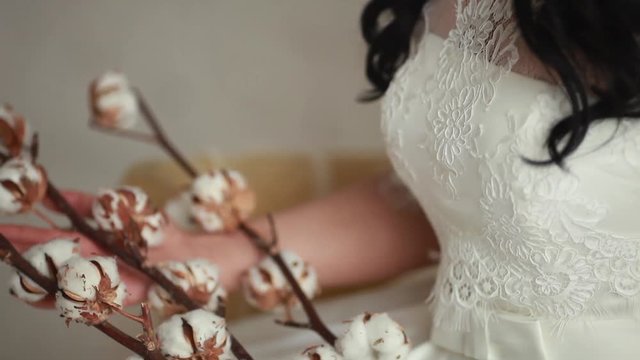 Tanned beautiful brunette bride in white wedding dress sitting on the chair. In the hands holding the cotton branches