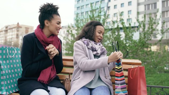 Two young african american women sharing their new purchases in shoppping bags with each other. Attractive girls talking while sitting on steet bench