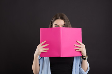 Fototapeta premium beautiful caucasian female student in denim shirt with long hair, over black background, covered her face with a book