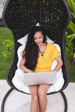 Young successful woman works as freelancer online in outdoor garden. Brunette in yellow dress with silver laptop. Internet work in travel