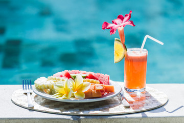 White plate with fresh fruit and juice with slice orange. Healthy breakfast on poolside in hotel in summertime. Plate with papaya, pineapple, watermelon, lime.