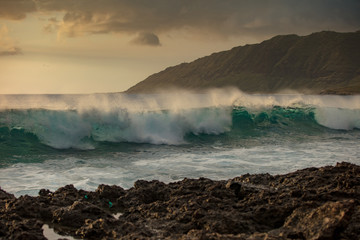 Beautiful green rough ocean wave incoming. Sunset sea with shorebreak water surface. Rocky coastline with mountain landscape on background