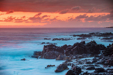 Beautiful red orange sunset in Pacific ocean. White splashes of ocean waves breaking against rocky coast.