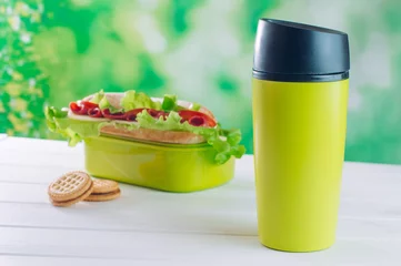 Gordijnen Green thermos mug near lunch box on white wooden background © lithiumphoto