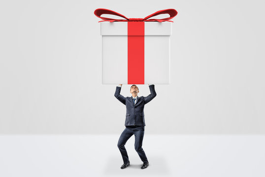 A small businessman holding a giant gift box with both hands above his head.