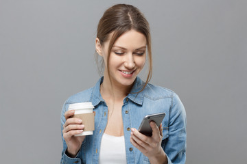 Closeup of young beautiful European lady isolated on gray background holding cardboard coffee cup and smartphone in her hands looking at screen and smiling happily at content she is watching.