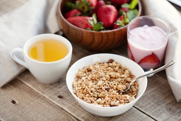 A bowl of homemade granola with yogurt and fresh strawberries on a wooden background. Healthy breakfast with green tea