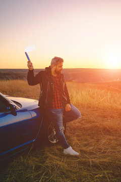 Young man holding burning torch in hand near his sports car at sunset