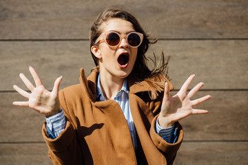 Fashion trendy casual young woman in sunglasses, brown coat showing shock emotion over brown wooden...