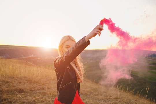 Happy woman enjoying nature sunset dressed in a red long dress and a black leather jacket holding smoke bomb