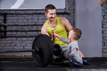 Young sporty father and little cute son sitting near barbell against brick wall in the cross fit...
