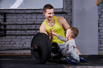 Young sporty father and little cute son sitting near barbell against brick wall in the cross fit gym.