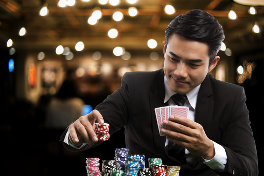 Portrait handsome man in black suit is putting stack of chips and holding poker card
