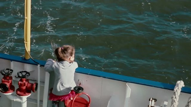 A little girl is standing closely to the board of a ship deck and looking at the water