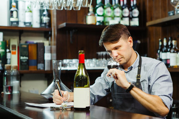 Male sommelier tasting red wine and making notes at bar counter