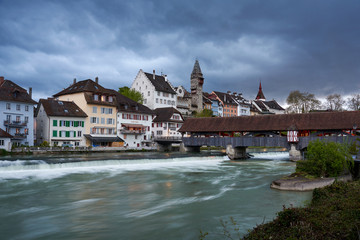 Obraz premium Regenwolken über Bremgarten, Holzbrücke über Fluss Reuss, verwischtes Wasser, Städtchen mit Turm, Kirche und Brücke